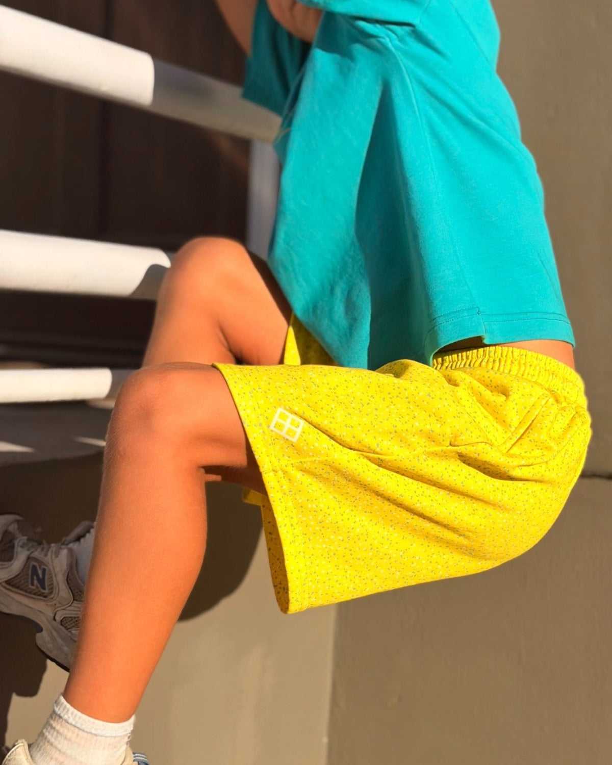 A child wearing bright yellow shorts and a turquoise t-shirt sits on outdoor steps, with sunlight casting strong shadows on a concrete wall.