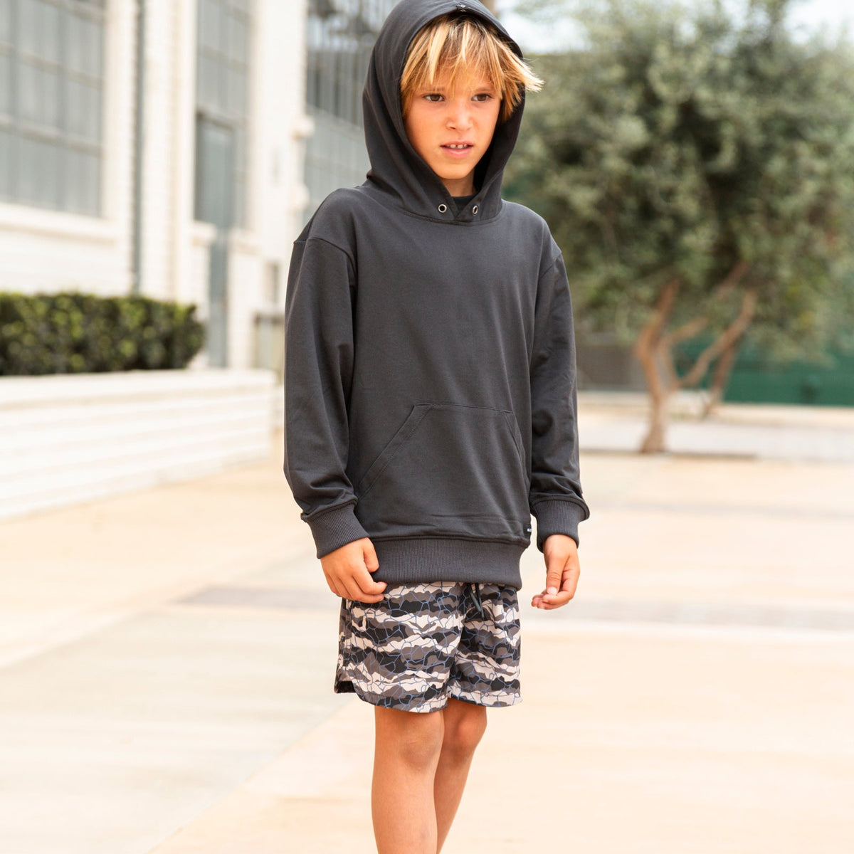 A young boy wearing a dark charcoal gray pullover hoodie, patterned shorts, white socks, and sneakers, standing outdoors on a paved walkway in front of a building.