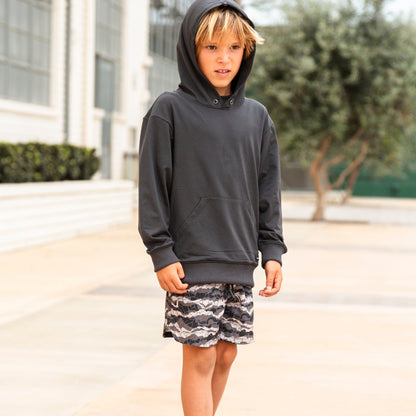 A young boy wearing a dark charcoal gray pullover hoodie, patterned shorts, white socks, and sneakers, standing outdoors on a paved walkway in front of a building.
