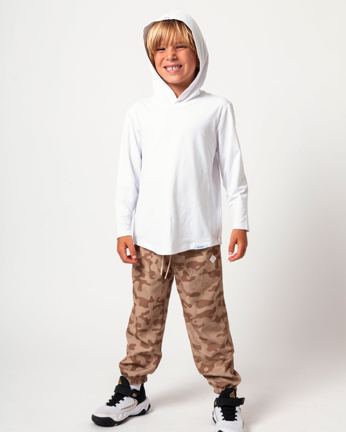A young boy wearing a white long-sleeve hooded shirt and brown camouflage jogger pants, standing against a plain white studio background.