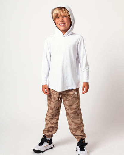 A young boy wearing a white long-sleeve hooded shirt and brown camouflage jogger pants, standing against a plain white studio background.