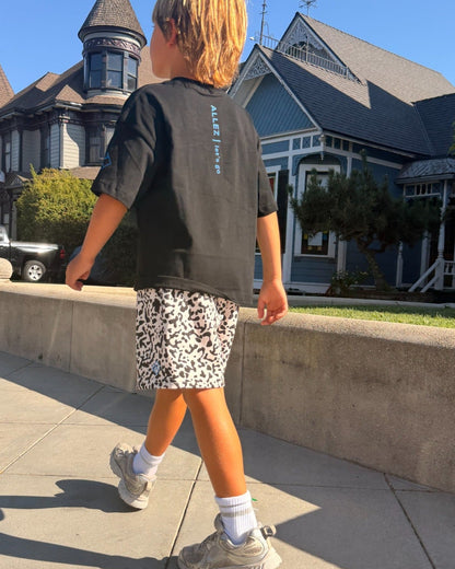 A child walking outdoors on a sunny day, viewed from behind. The child is wearing a black t-shirt with 'AESTHETE' printed vertically on the back, patterned shorts, white socks, and gray sneakers, with a large Victorian-style house in the background.