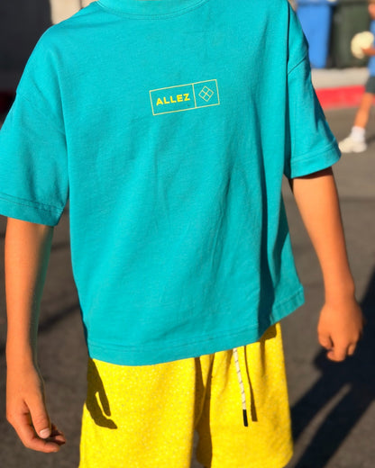 A child wearing a bright turquoise short-sleeve t-shirt with a small yellow and white 'ALLEZ' graphic on the chest, paired with yellow shorts, photographed outdoors in bright sunlight.