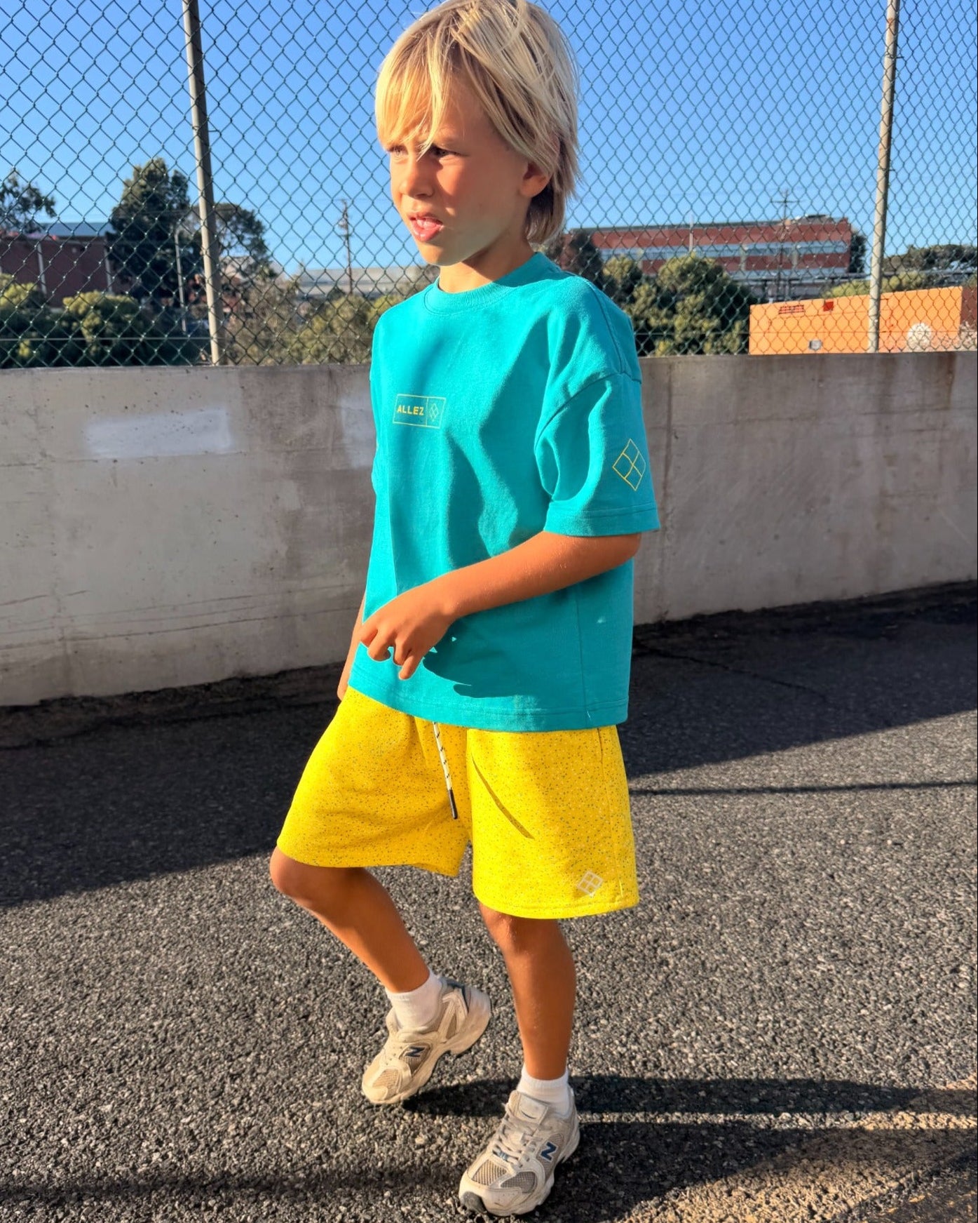 A young blonde boy wearing a bright turquoise t-shirt and yellow shorts stands on an outdoor paved surface near a chain-link fence.