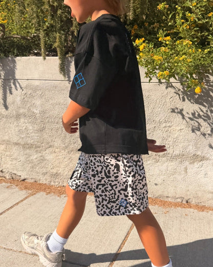 A young boy walking outdoors on a sunny sidewalk, wearing a black t-shirt with a small blue geometric logo, black and white leopard-print shorts, white socks, and gray sneakers.