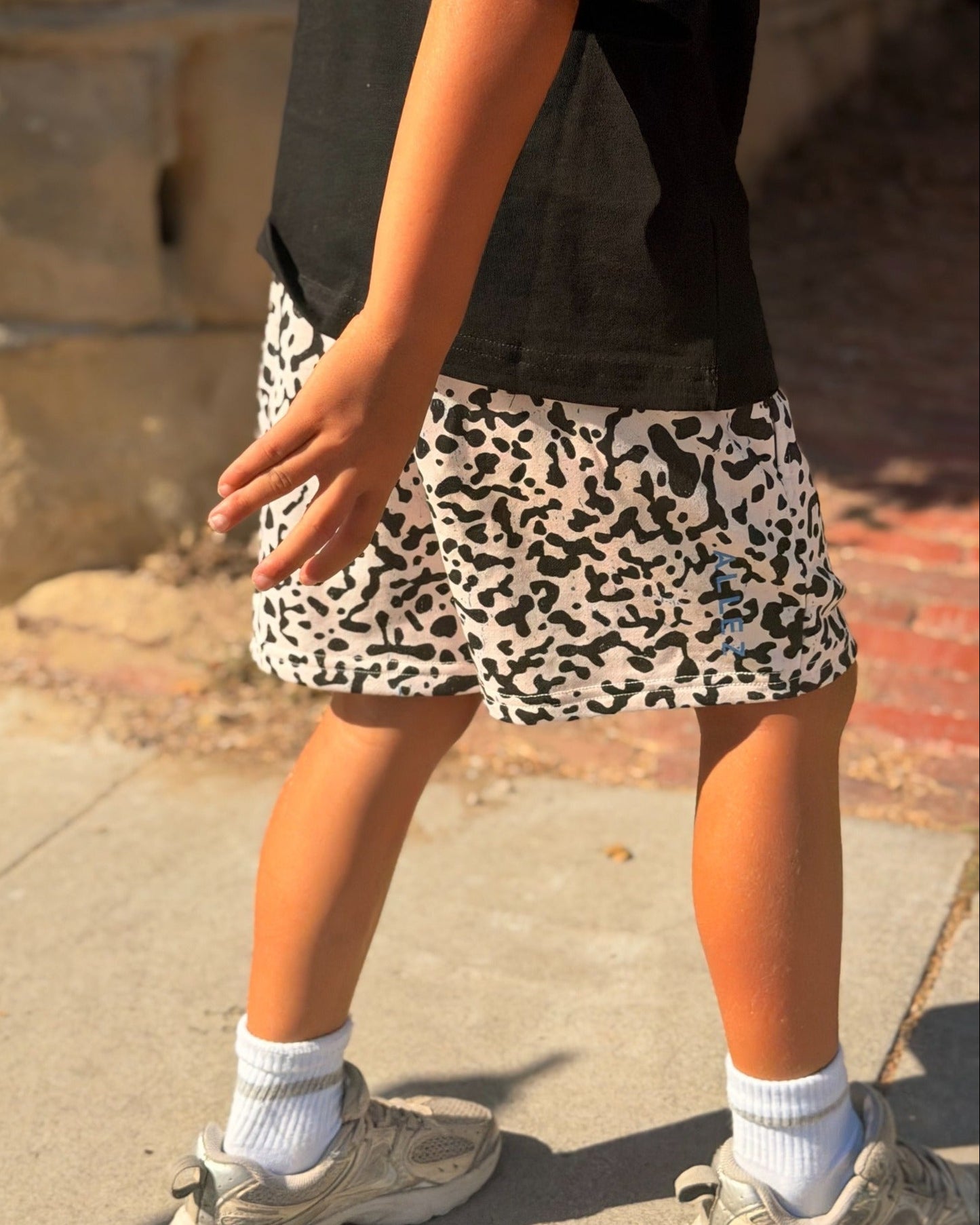 A child wearing a black t-shirt and a black and white leopard print shorts standing outdoors on a concrete sidewalk.