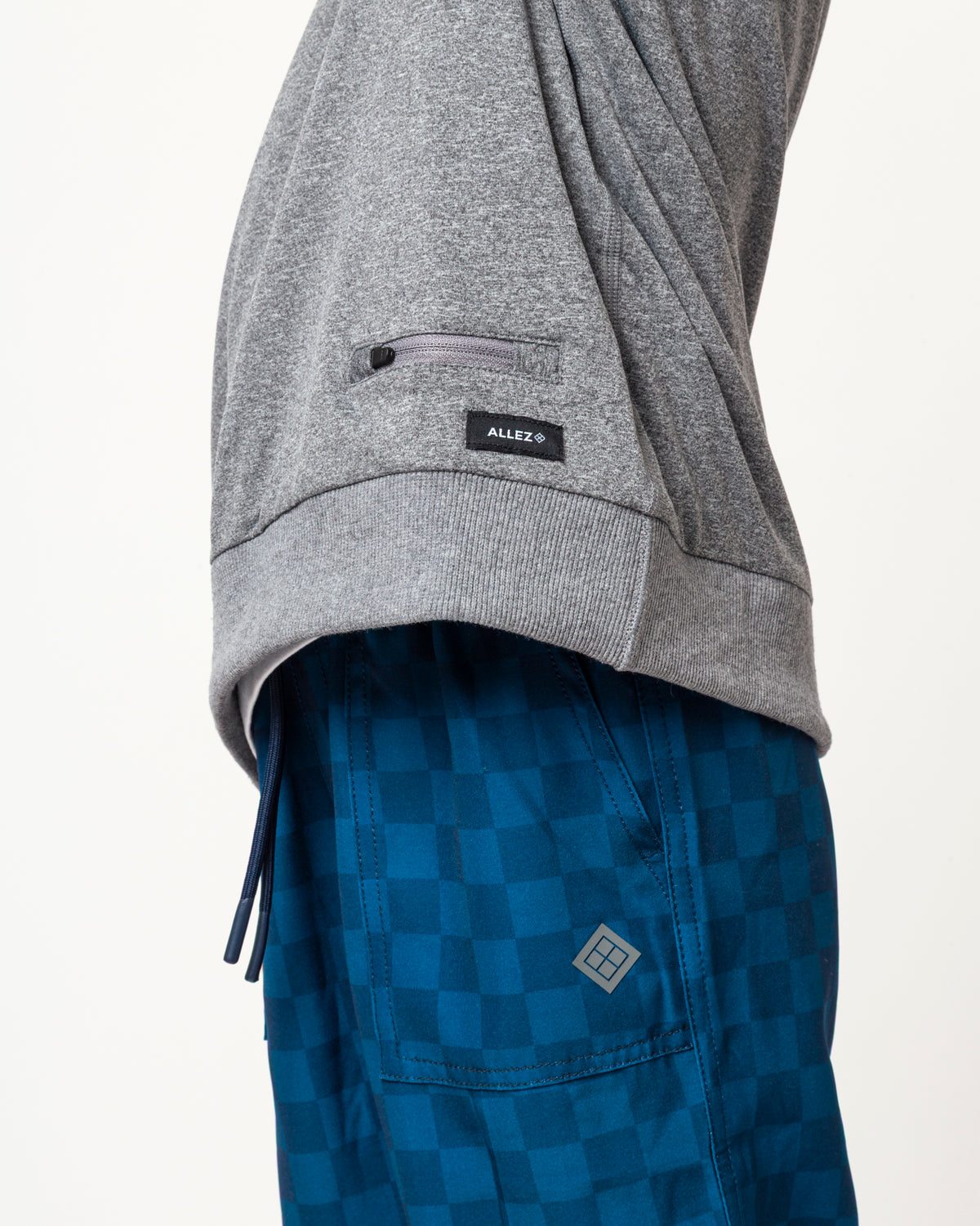 Close-up of a person wearing a heather gray zippered pocket sweatshirt and blue checkered board shorts, showing a small black Alex's brand patch on the sleeve and a matching logo patch on the shorts.
