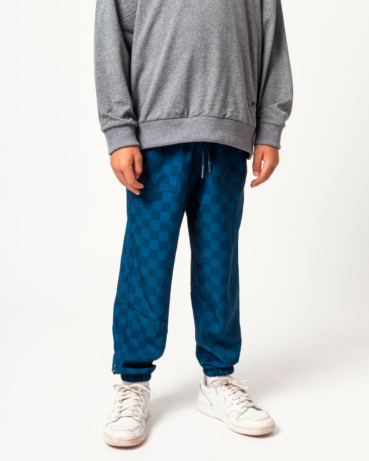 A person wearing blue checkered jogger pants, a gray sweatshirt, and white sneakers, standing against a plain white studio background.
