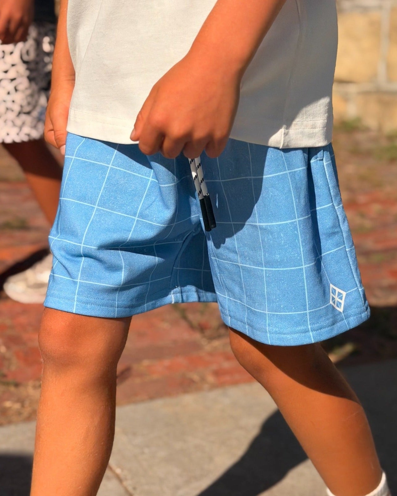 A child wearing light blue grid-patterned shorts, a white t-shirt, white socks, and New Balance sneakers walking outdoors on a paved path.