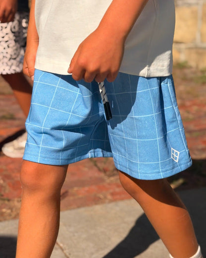 A child wearing light blue grid-patterned shorts, a white t-shirt, white socks, and New Balance sneakers walking outdoors on a paved path.