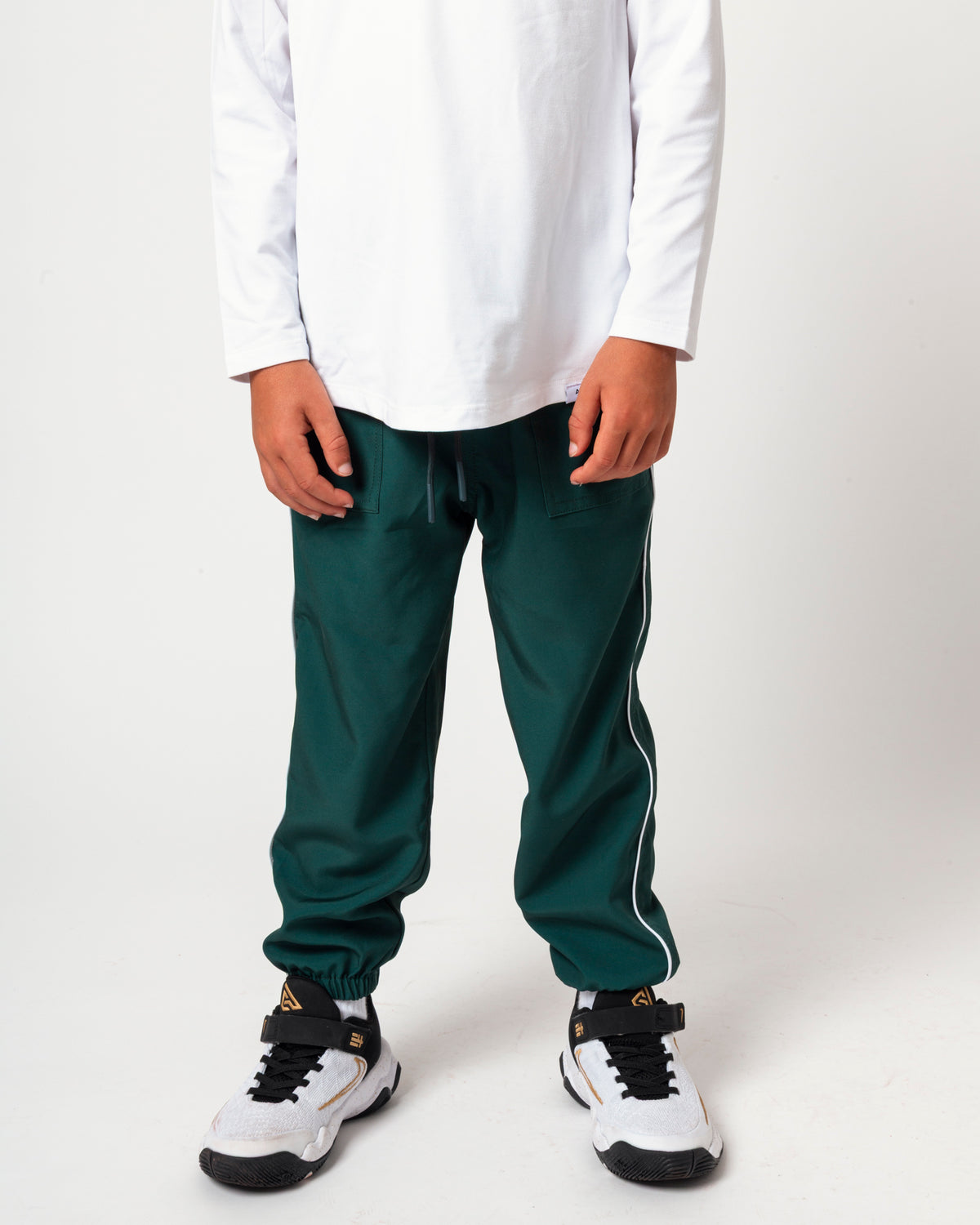 A child wearing dark green jogger-style pants with white side stripes, a white long-sleeve shirt, and white sneakers with black details, standing against a light gray studio background.