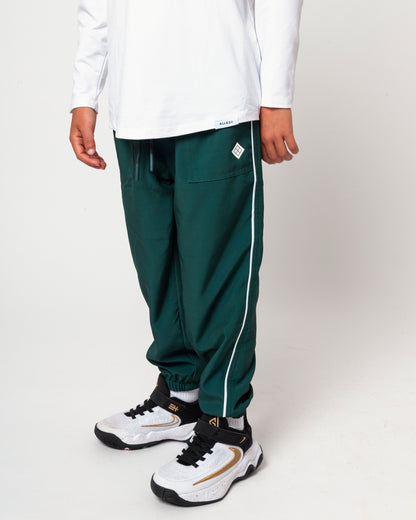 A person wearing dark green jogger-style pants with a white side stripe, a white long-sleeve top, and white sneakers with black and gold details, photographed against a light gray studio background.